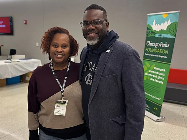 Kobieta z identyfikatorem konferencyjnym stoi obok uśmiechniętego mężczyzny w okularach i szarej marynarce, na tle banera z napisem „Chicago Parks Foundation, Where park users and lovers become park givers and stewards”.