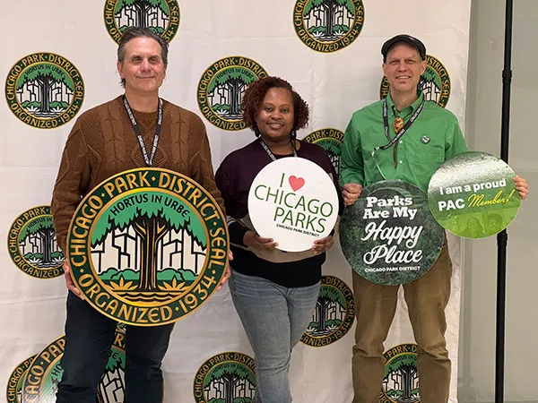 Trzy osoby pozują na tle ścianki z nadrukowaną pieczęcią Chicago Park District. Jedna trzyma dużą pieczęć z napisem „Chicago Park District Seal, Hortus in Urbe, Organized 1934”; pozostałe trzymają okrągłe tabliczki z napisami „I love Chicago Parks”, „Parks Are My Happy Place” i „I am a proud PAC Member!”.