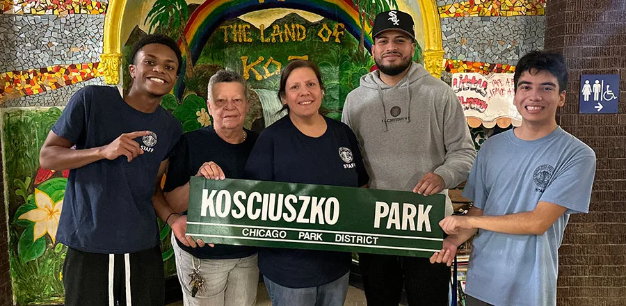 Pięć osób, w tym cztery w koszulkach pracowników parku, trzyma zielony szyld z napisem „Kosciuszko Park, Chicago Park District” na tle mozaikowego muralu z tęczą i słowami „The Land of Koz”.